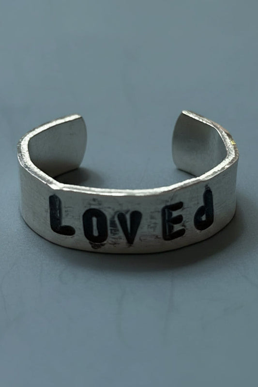 Silver colored ring with 'LOVED' engraving by hand stamped on a light gray background.