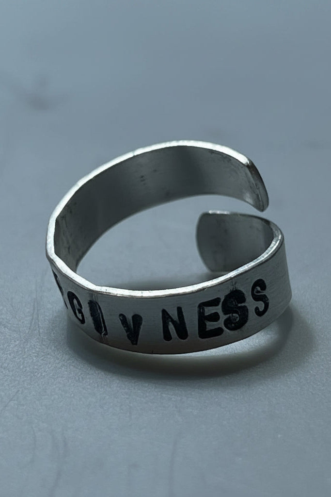 Silver color ring with 'FORGIVENESS' engraved by hand stamping on a gray surface.