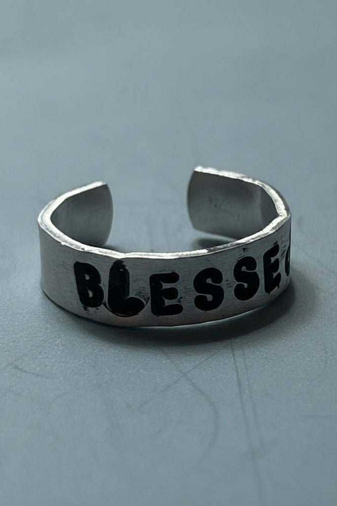 Silver colored ring with 'BLESSED' engraved by hand stamping on a light gray surface.