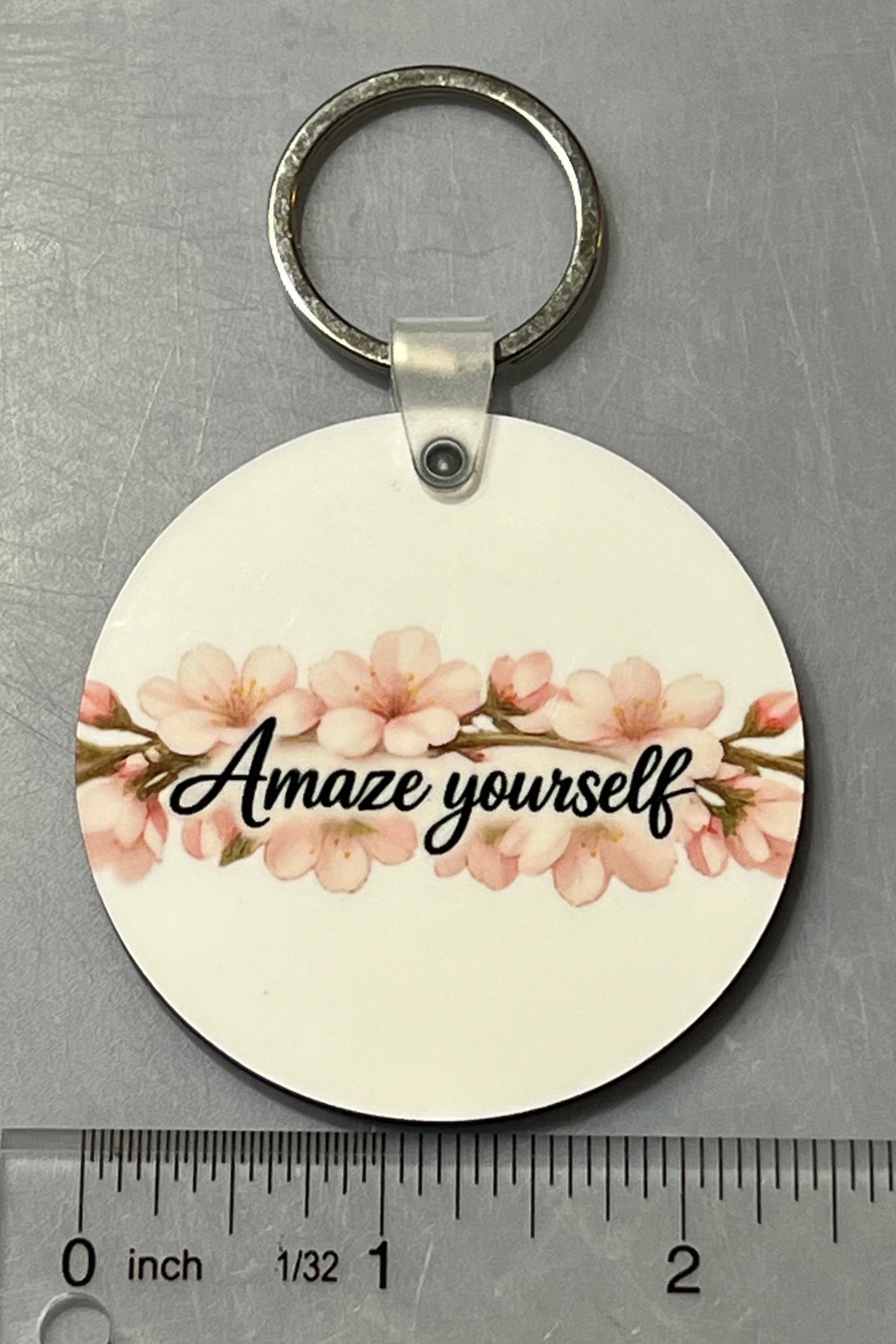 A round wood keychain with a silver split ring, featuring a white background, pink cherry blossoms image and the text 'Amaze yourself' in black script. A clear plastic ruler is positioned below the keychain to show its size against a gray metal background.