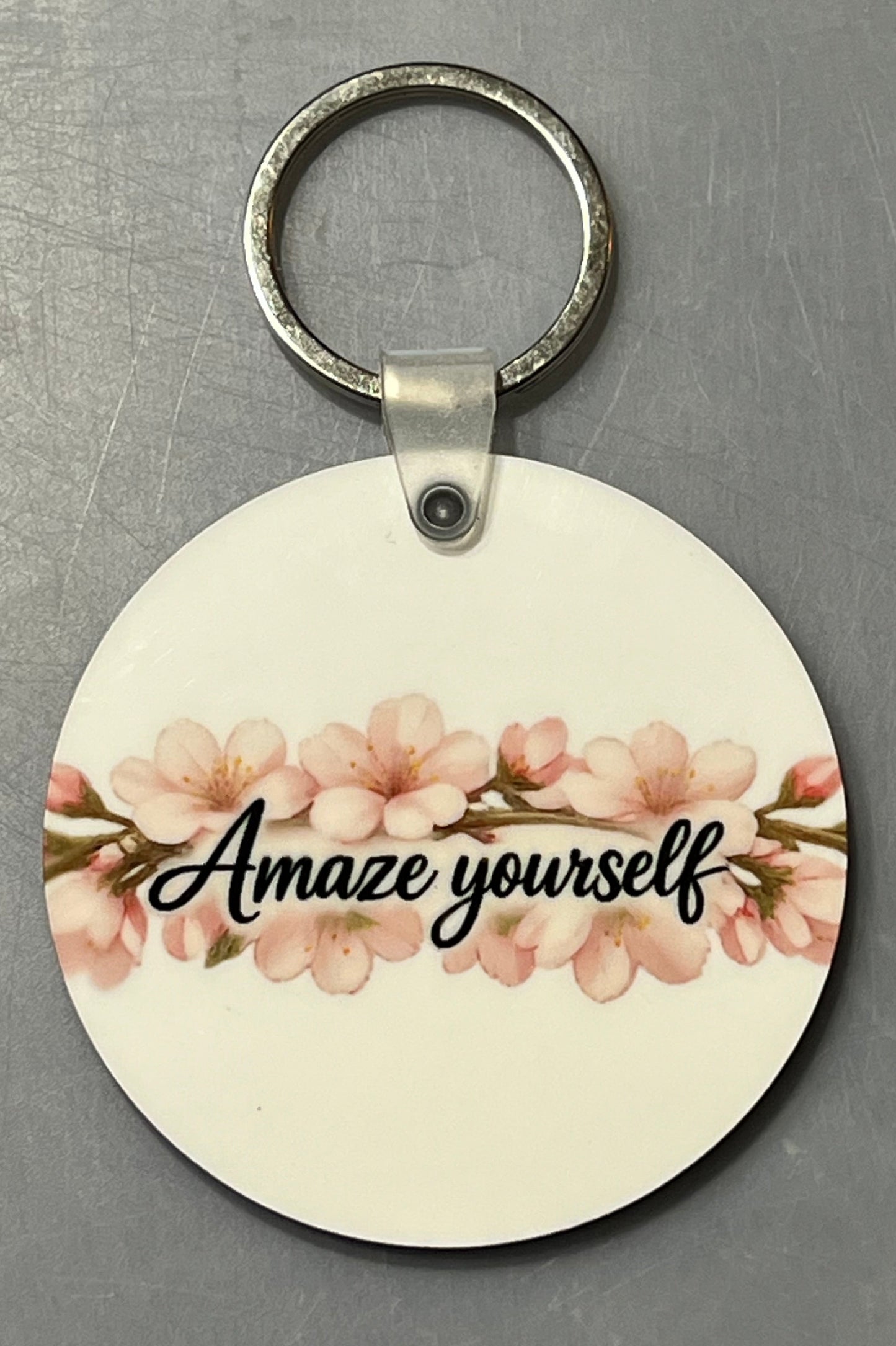 A round white wooden keychain featuring a pink cherry blossom design and the text 'Amaze yourself' in black script, attached to a silver metal key ring, set against a textured gray background.