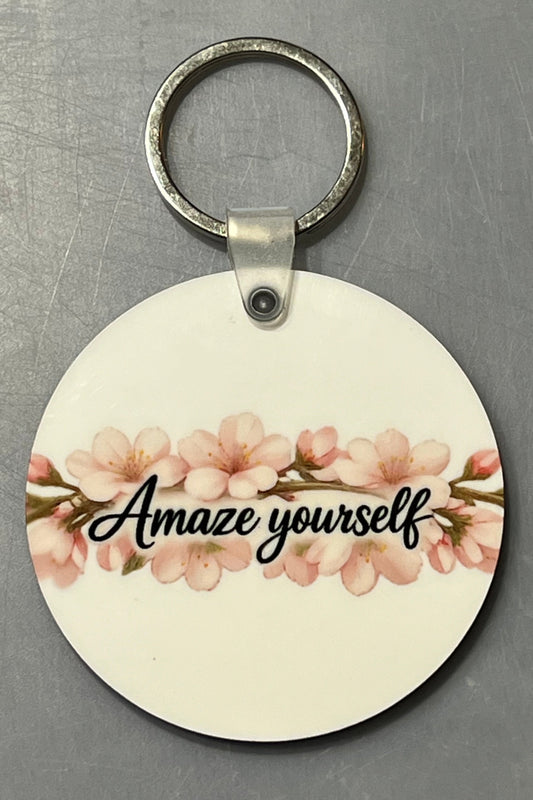 A round white wooden keychain featuring a pink cherry blossom design and the text 'Amaze yourself' in black script, attached to a silver metal key ring, set against a textured gray background.
