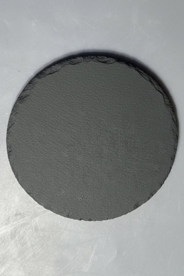 A round dark gray slate piece with natural rough edges, shown on a blue-gray surface.