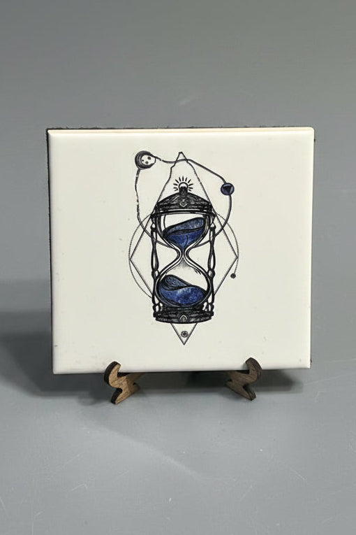 A square white ceramic tile plaque featuring a black line-art illustration of an hourglass with blue stone-like centers, photographed on a gray surface with small wooden supports visible on the right.