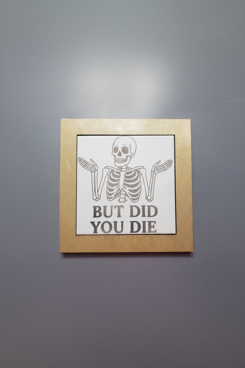 Trivet hot plate with wood frame around a ceramic tile.  Shows image of a skeleton shrugging with his hands raised saying 'But Did You Die'