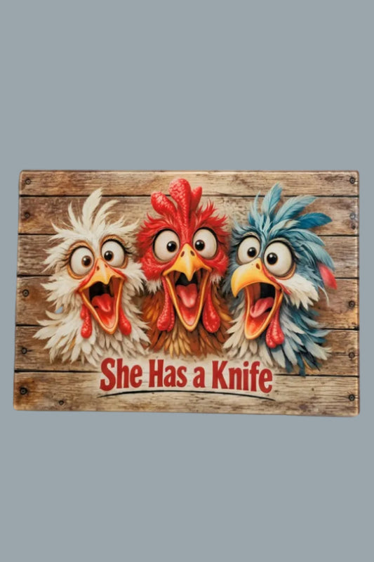 A rectangular rustic style glass cutting board featuring three cartoon chickens with wide-open mouths and the text 'She Has a Knife' in red at the bottom, shown against a plain light background.