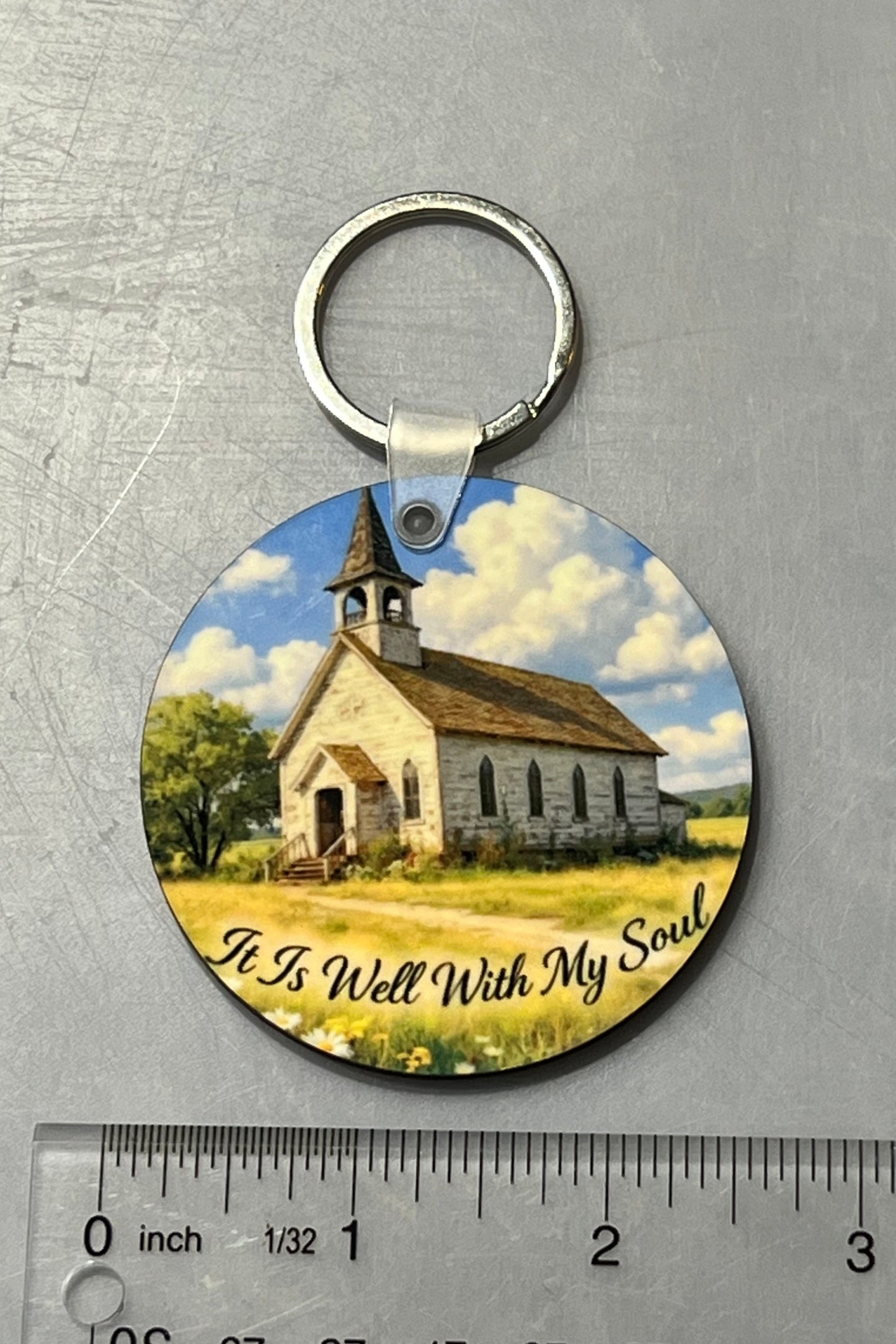 A round wood keychain featuring a printed illustration of a small church in a grassy field under a blue sky, with the text 'It Is Well With My Soul' along the bottom. The keychain is displayed on a gray textured surface above a ruler for scale.