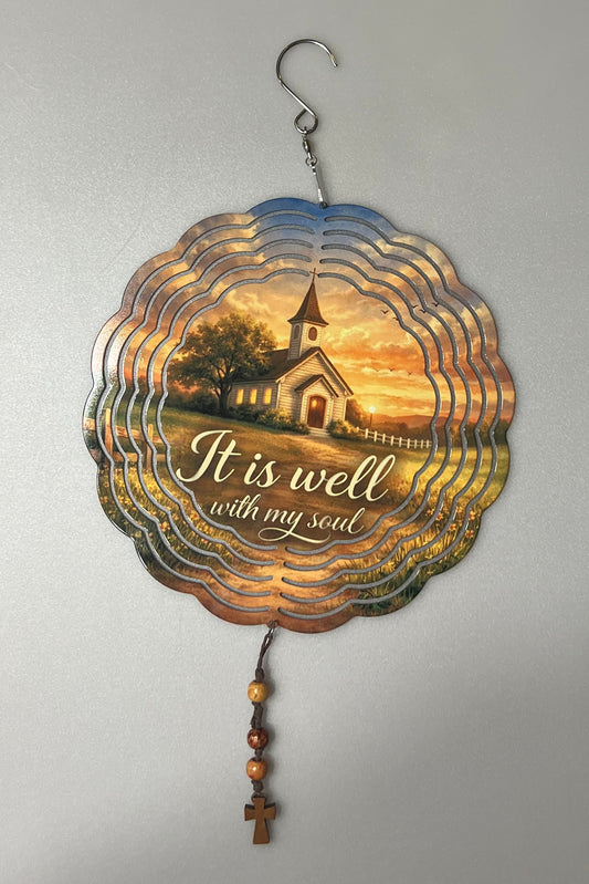 A scalloped metal wind spinner ornament with a rustic sunset design, featuring the text 'It is well with my soul' and a small hanging hook on one side and a beaded string on the other.