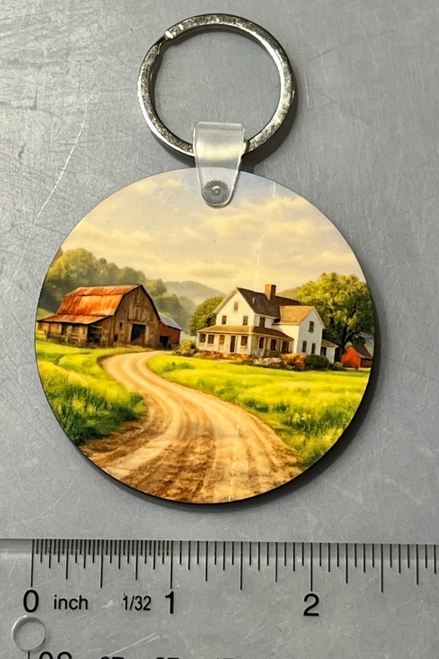 A round wooden keychain featuring a printed scenic landscape of a farm road with houses and barns, shown on a gray textured background with a clear ruler below for scale.