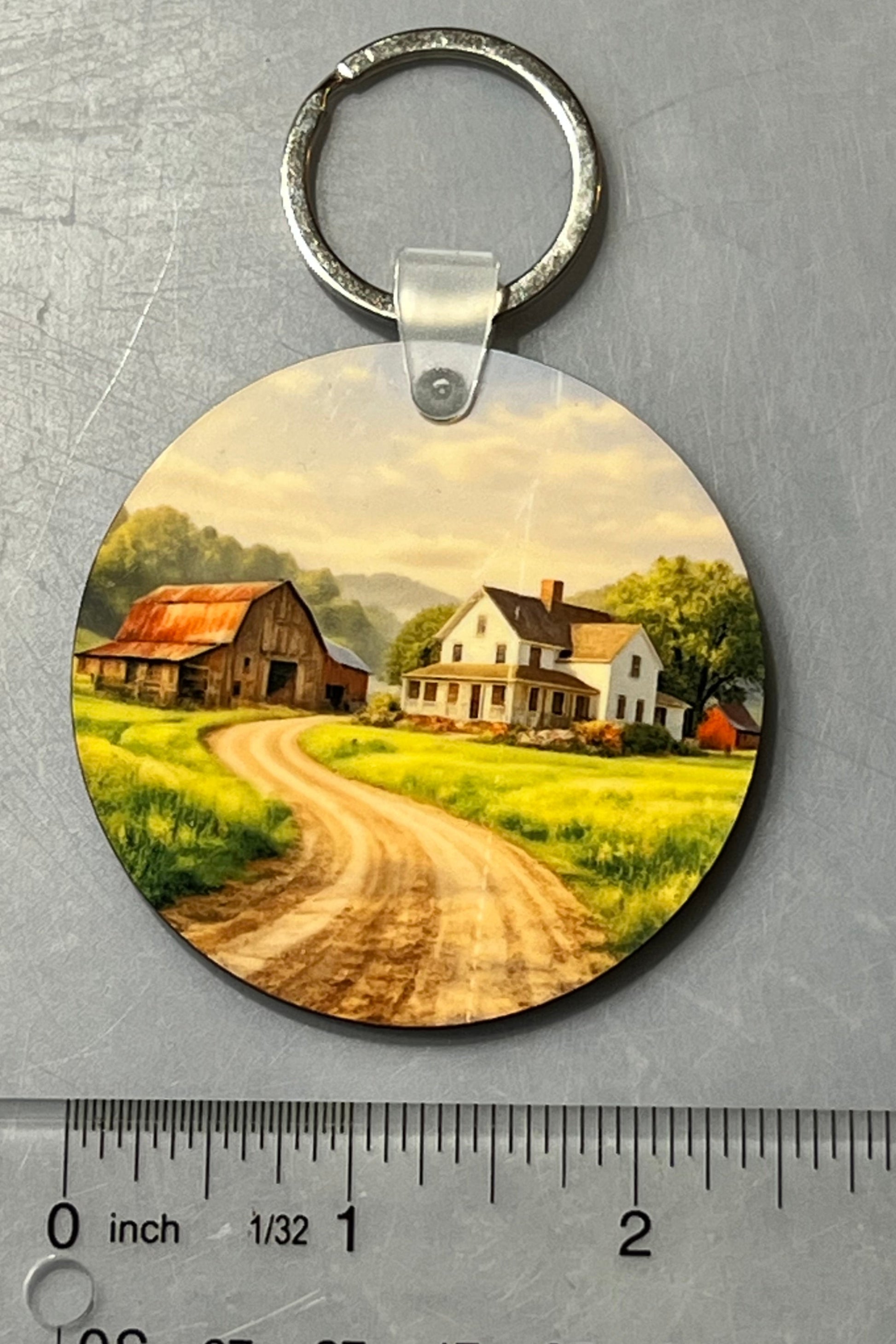 A round wooden keychain featuring a printed scenic landscape of a farm road with houses and barns, shown on a gray textured background with a clear ruler below for scale.