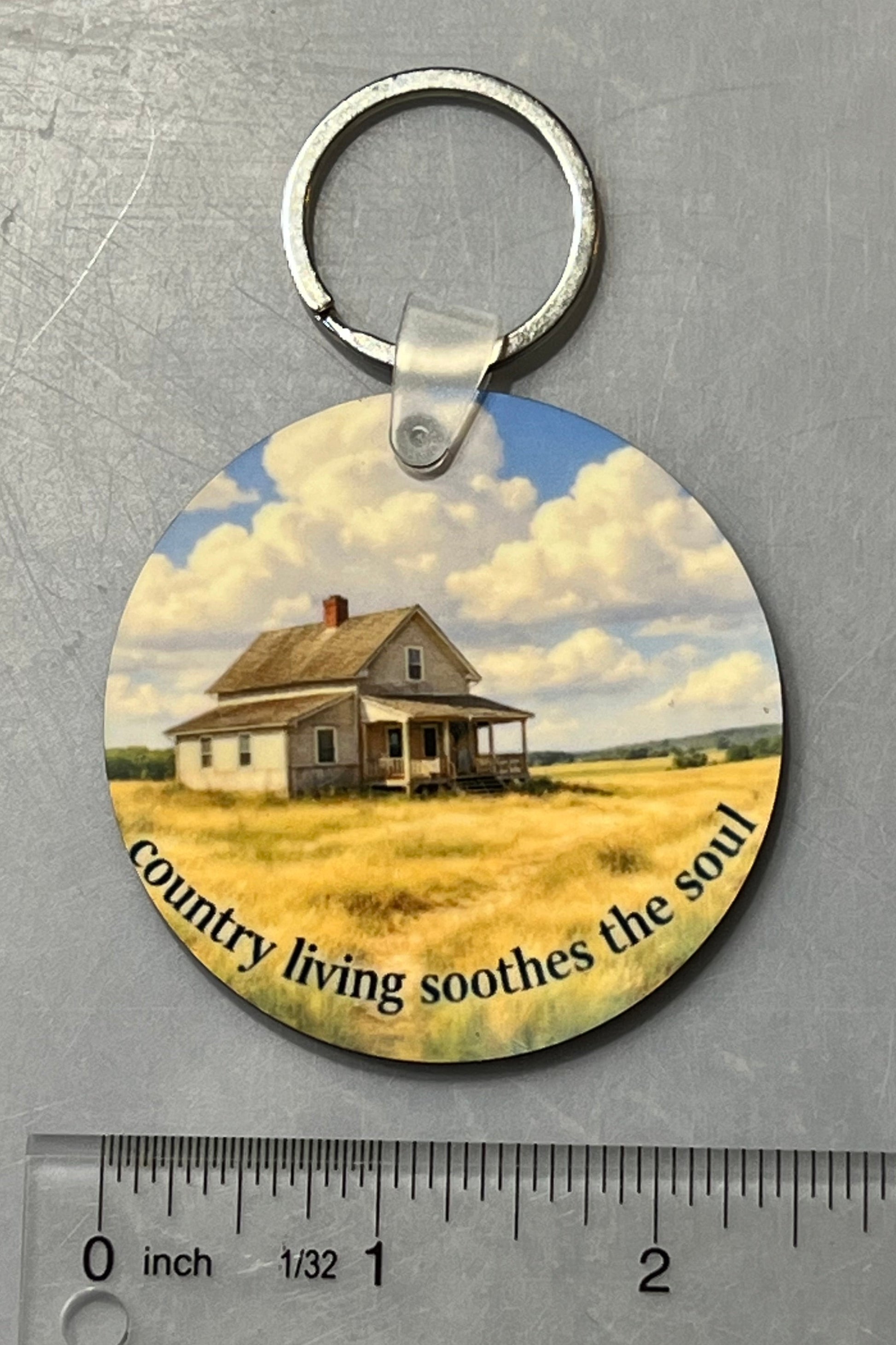 A round printed keychain with a silver metal key ring, featuring an illustration of a small house in a field under a cloudy blue sky, with the text 'Country living soothes the soul' around the bottom edge. A clear ruler is placed below it on a gray metal surface to show size.