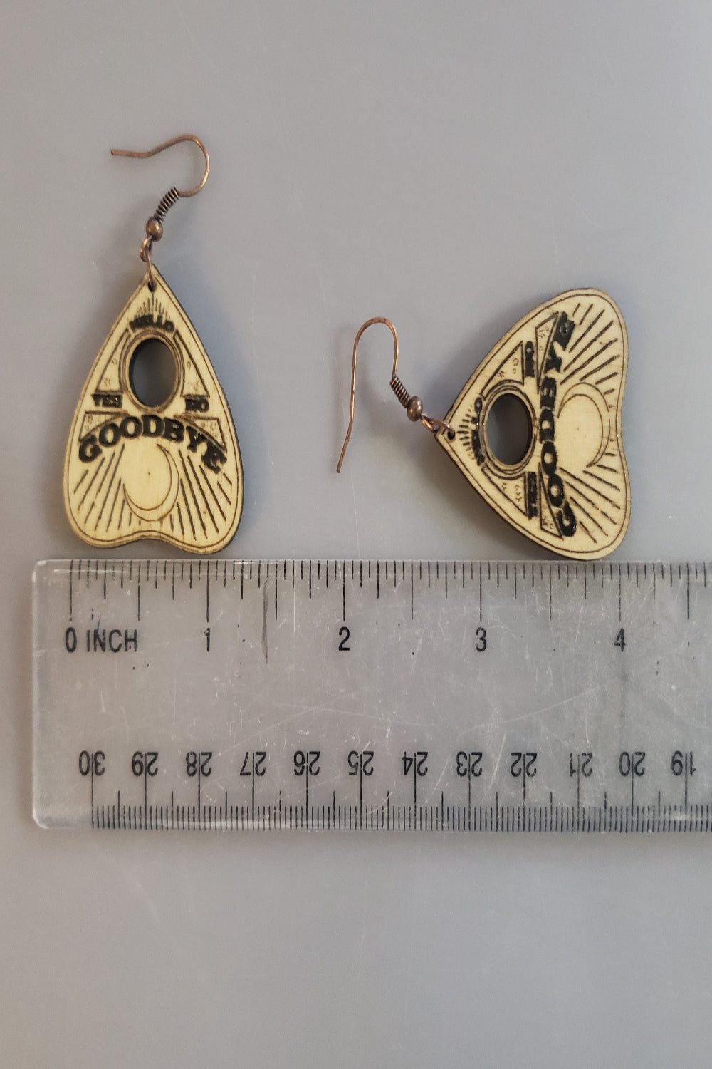 Wood handel heart-shaped earrings with 'Goodbye' text next to a ruler for scale on a gray background