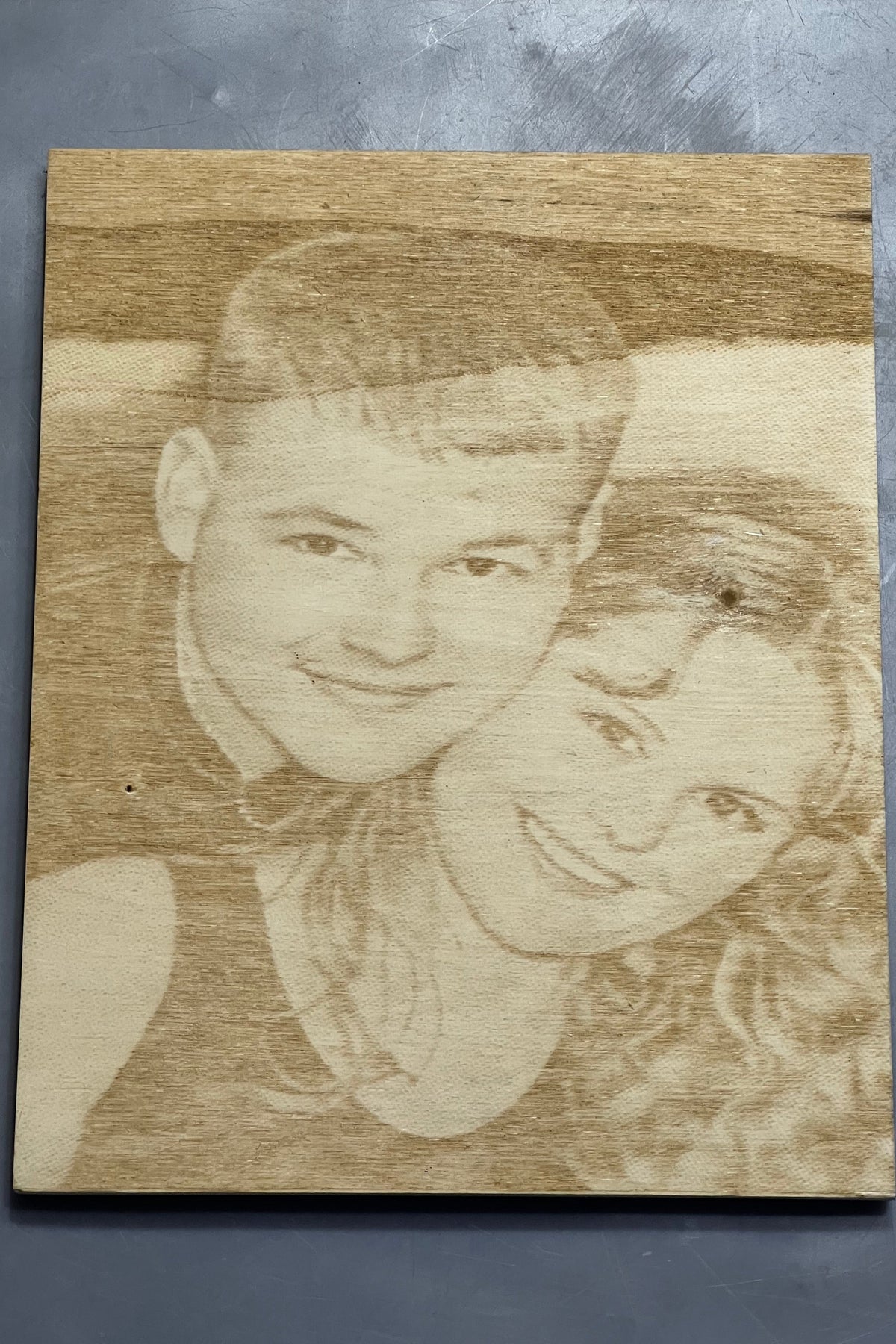 A rectangular wooden plaque featuring a custom laser-engraved portrait of a couple, displayed against a textured dark gray background.