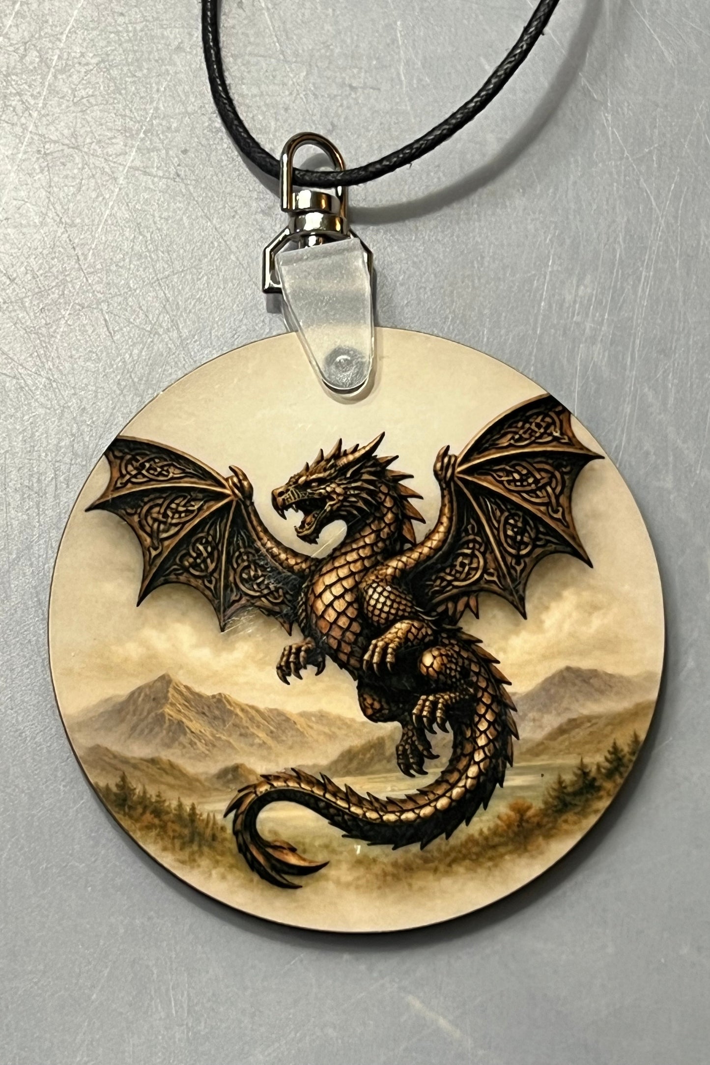 A round wooden pendant necklace featuring a detailed illustration of a bronze dragon with spread wings over a mountain landscape, suspended from a black cord on a textured gray background.