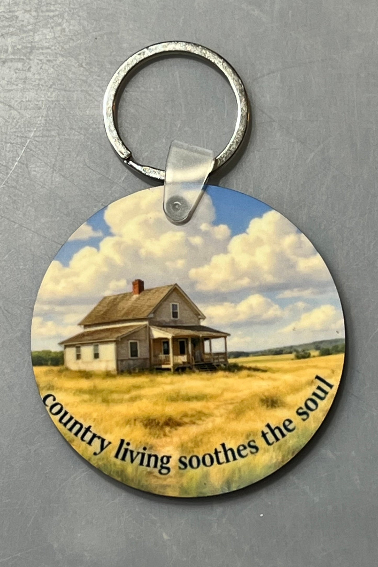 A round wood keychain featuring a printed scenic illustration of a small farmhouse in a field under a cloudy blue sky, with the text 'country living soothes the soul' along the bottom. The keychain is photographed on a textured gray background.
