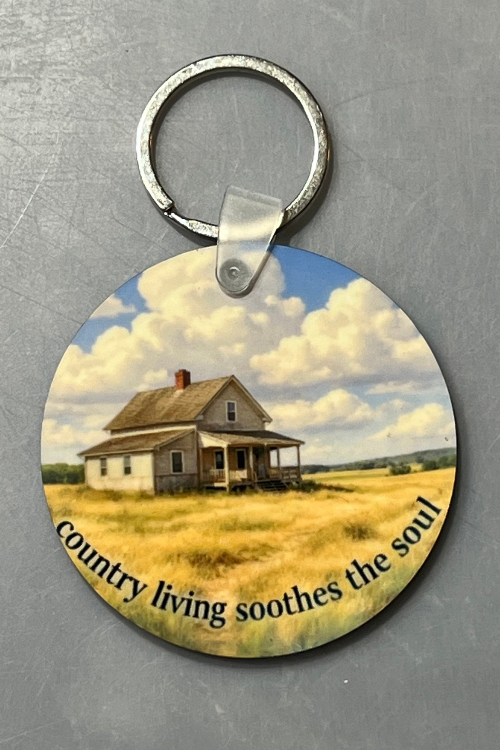 A round wood keychain featuring a printed scenic illustration of a small farmhouse in a field under a cloudy blue sky, with the text 'country living soothes the soul' along the bottom. The keychain is photographed on a textured gray background.
