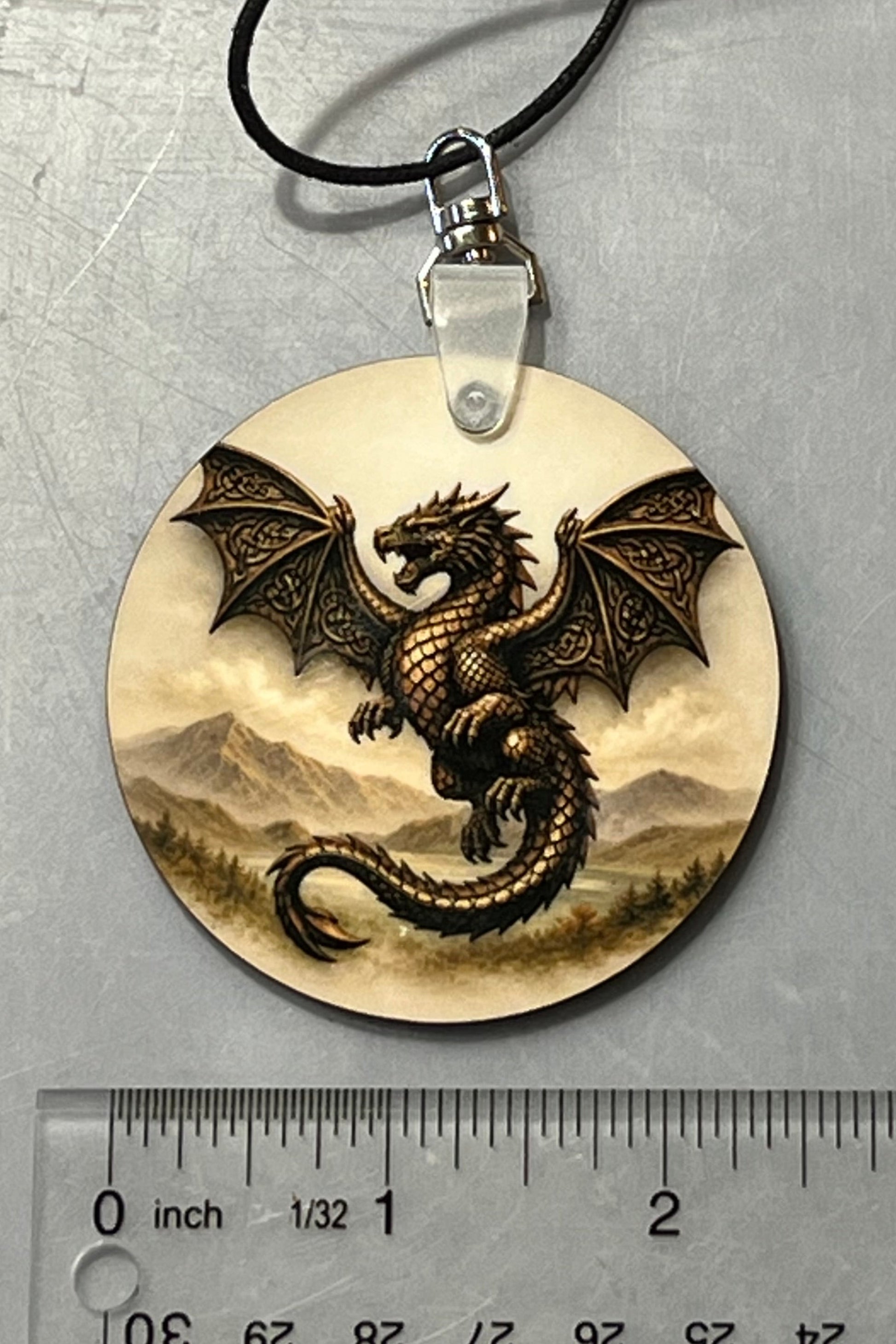 A round dangle featuring a detailed  dragon over a mountain landscape, suspended from a black cord with a swivel silver clasp, shown next to a ruler for scale on a gray background.