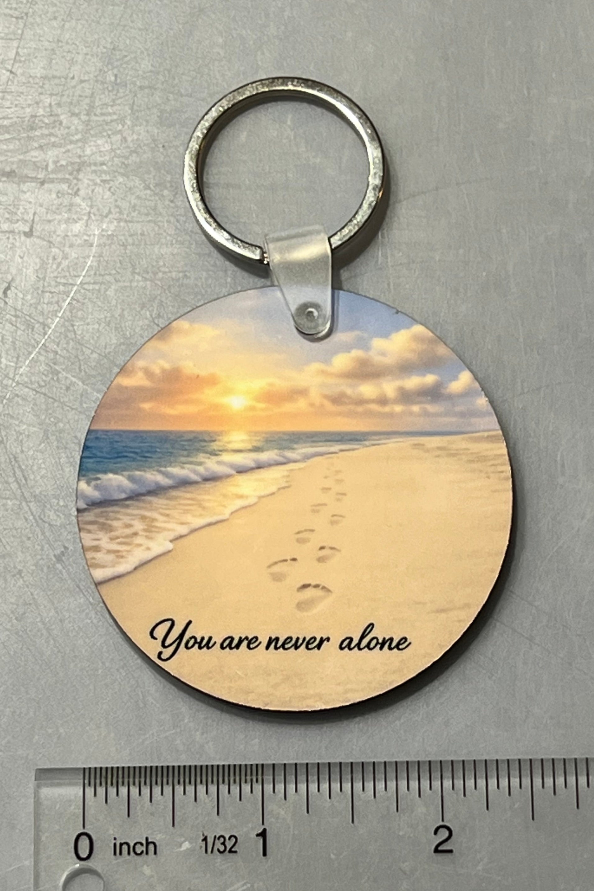 A round wood keychain featuring a beach sunset image with the text 'You are never alone', shown on a gray textured surface with a ruler below for scale. 2 1/2" Round.