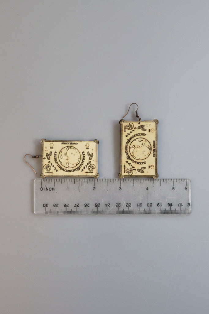 Spirit board shaped earrings with intricate designs on a ruler for scale.d