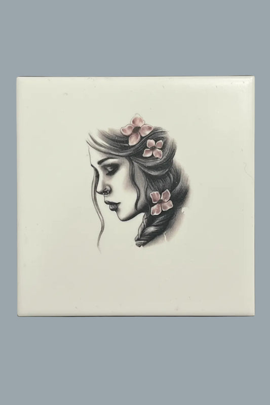 A square ceramic tile featuring a black and white sketch of a woman's profile with pink flowers in her hair.