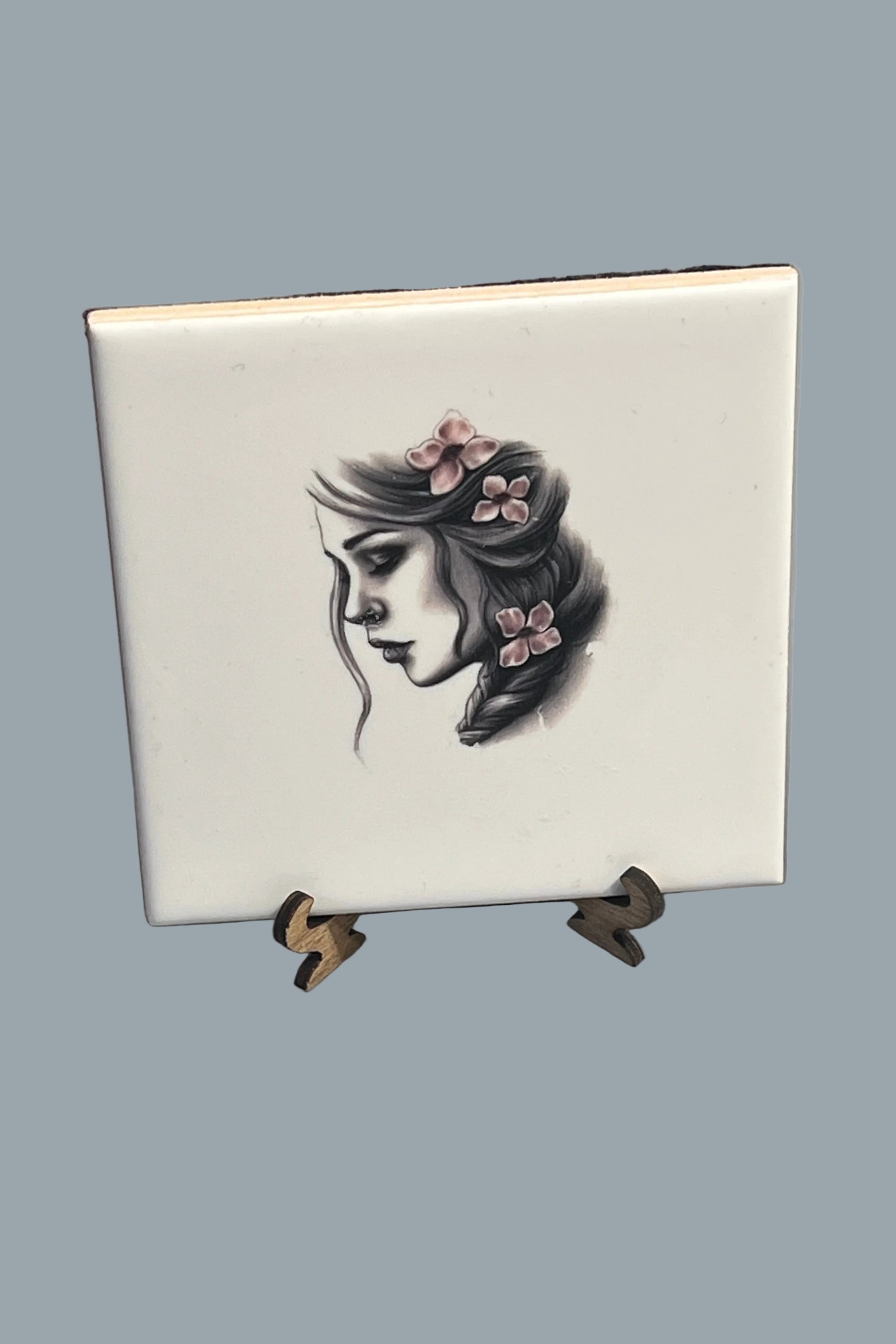 A square white ceramic plaque featuring a black ink illustration of a woman's face with pink flowers in her hair.
