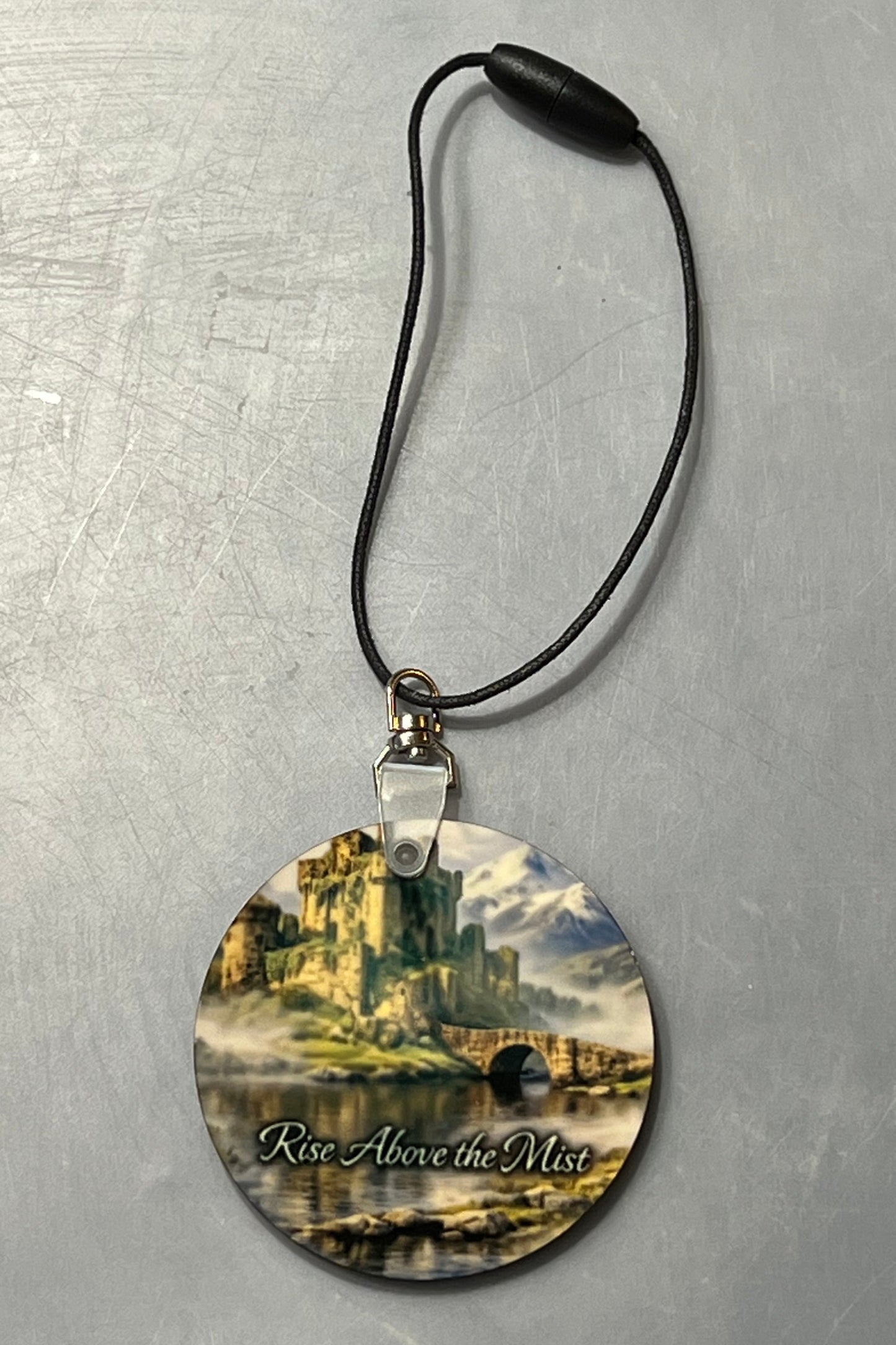 A circular decoration featuring a scenic fantasy castle illustration with the text 'Rise Above the Mist' on a black cord, displayed on a textured gray background.