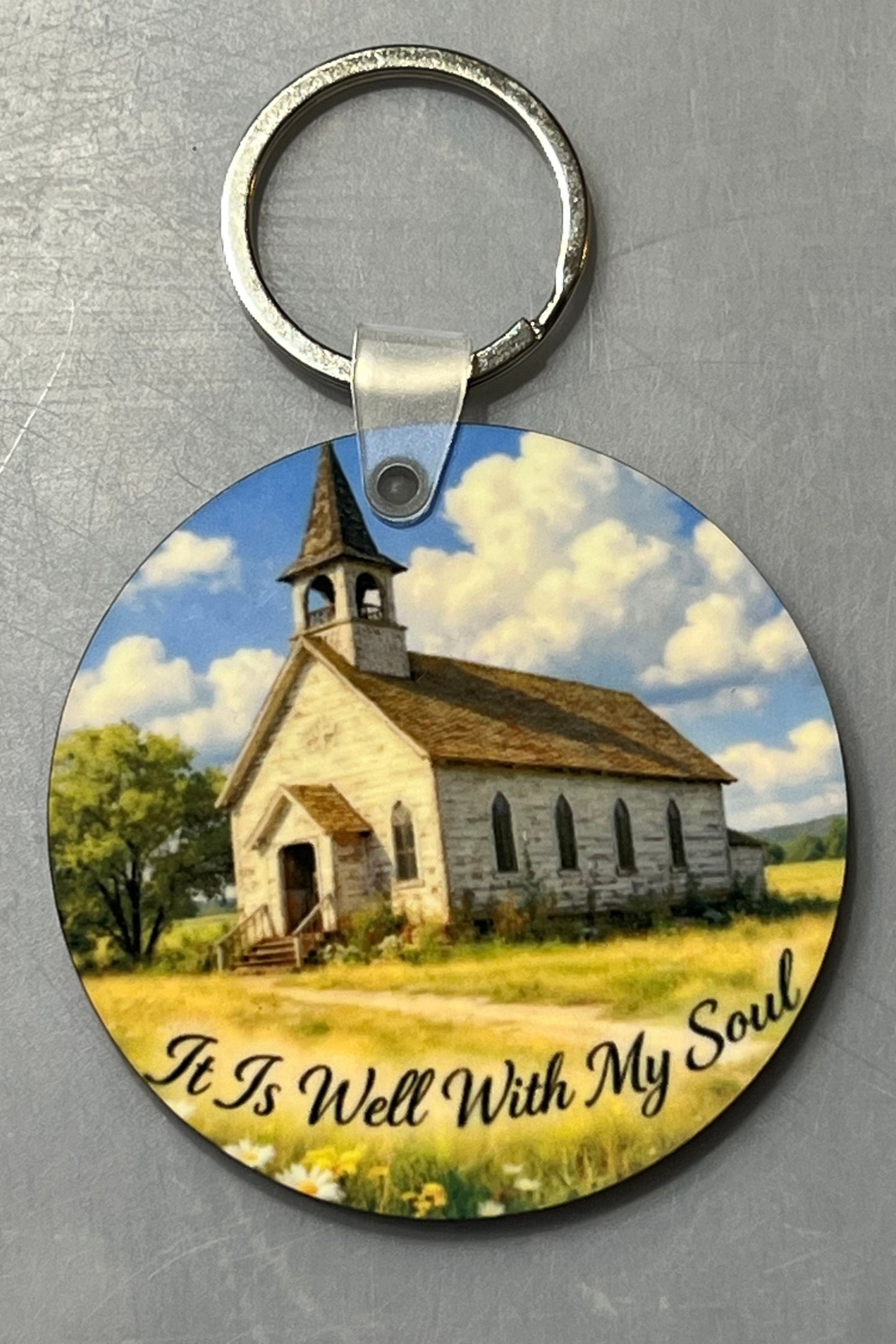 A round wood keychain featuring a printed scenic illustration of a small church in a grassy field under a blue sky with clouds, with the text 'It Is Well With My Soul' along the bottom. The keychain is photographed on a textured gray background.