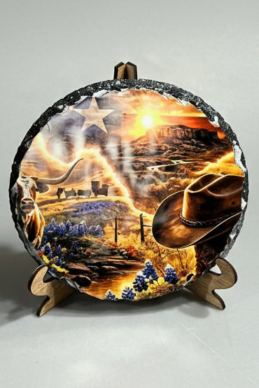 A round decorative stone plaque with a rustic black textured rim, displayed on a small wooden stand. The slate stone features a vibrant, detailed western landscape print with a longhorn steer, cowboy hat, bluebonnets, a covered wagon, and a sunset over mountains, alongside a white star and a stylized Texas flag.