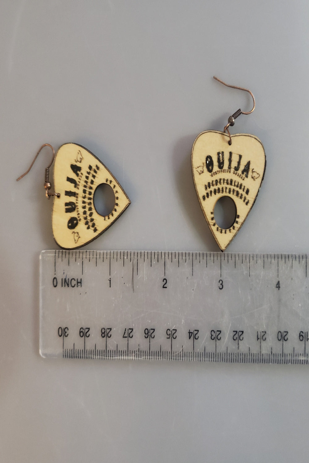 Heart-shaped Ouija board earrings with a ruler for scale on a gray background