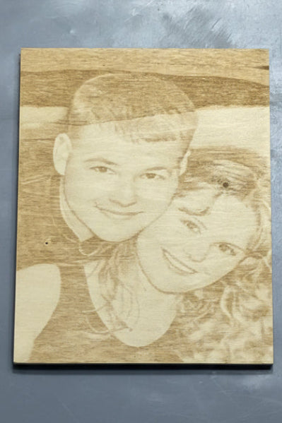 A rectangular wooden plaque featuring a laser-engraved portrait of two smiling people, shown against a textured gray background.