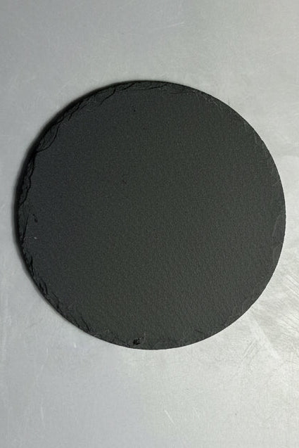 A round gray stone or slate with rough, natural edges, photographed from directly above on a light gray textured surface.