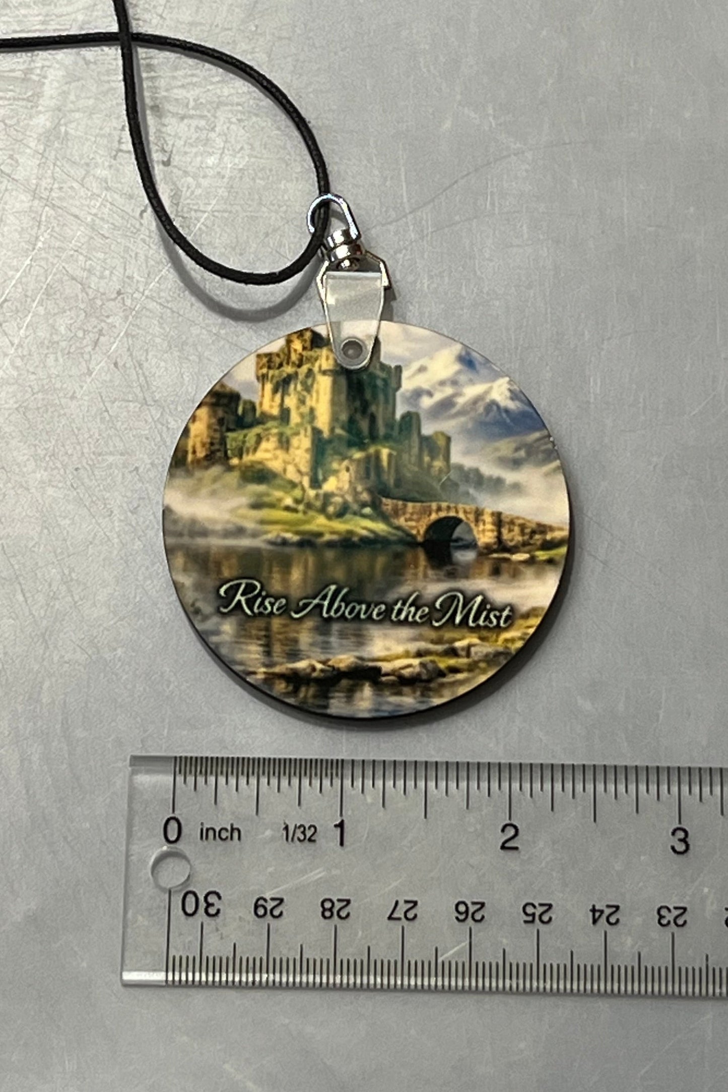 A round mirror hanger for your cars' interior, or anywhere you'd like to hang this inspirational message  featuring a scenic castle illustration and the text 'Rise Above the Mist,' suspended on a black cord with a ruler below for scale.