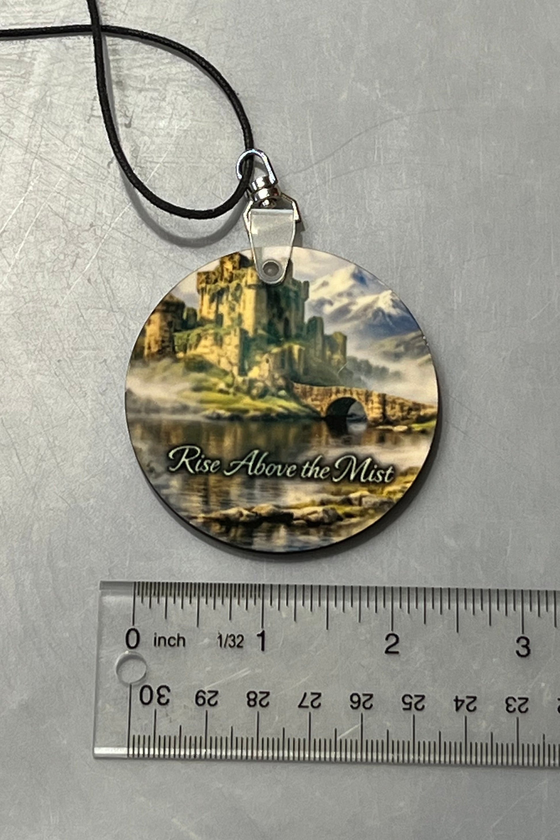 A round mirror hanger for your cars' interior, or anywhere you'd like to hang this inspirational message  featuring a scenic castle illustration and the text 'Rise Above the Mist,' suspended on a black cord with a ruler below for scale.