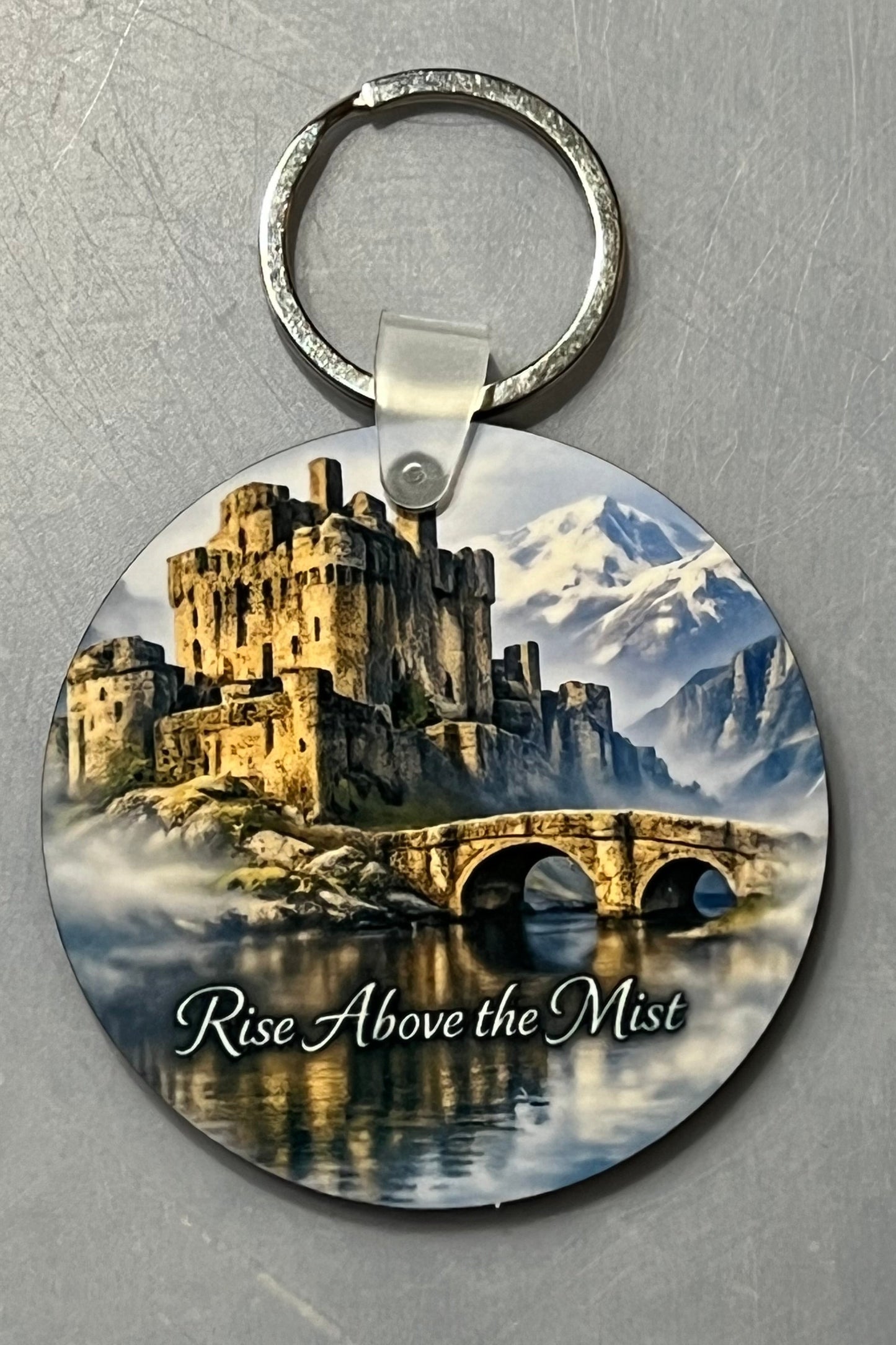 A round wood keychain featuring a scenic illustration of a stone castle, bridge, mountains, and misty water, with the text 'Rise Above the Mist' on a silver metal ring, set against a textured gray background.