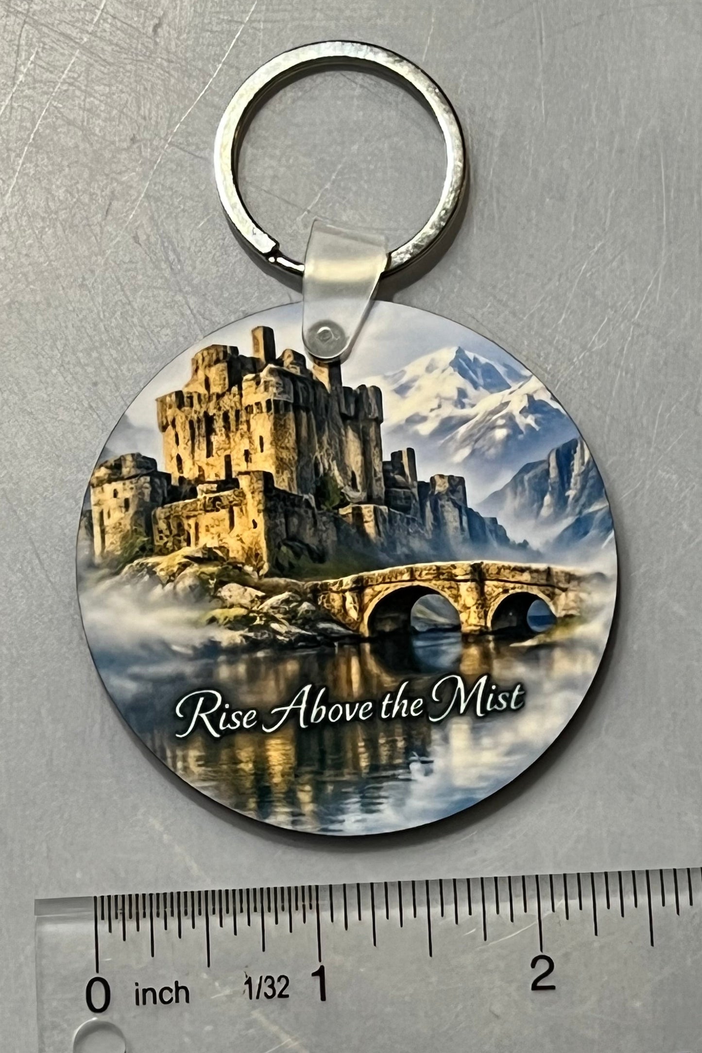 Stone castle with bridge in the foreground with mist rising above the water. The background shows a line of mountains in the distance.  Keychain is shown with a ruler for measure and is 2 1/2” round. 