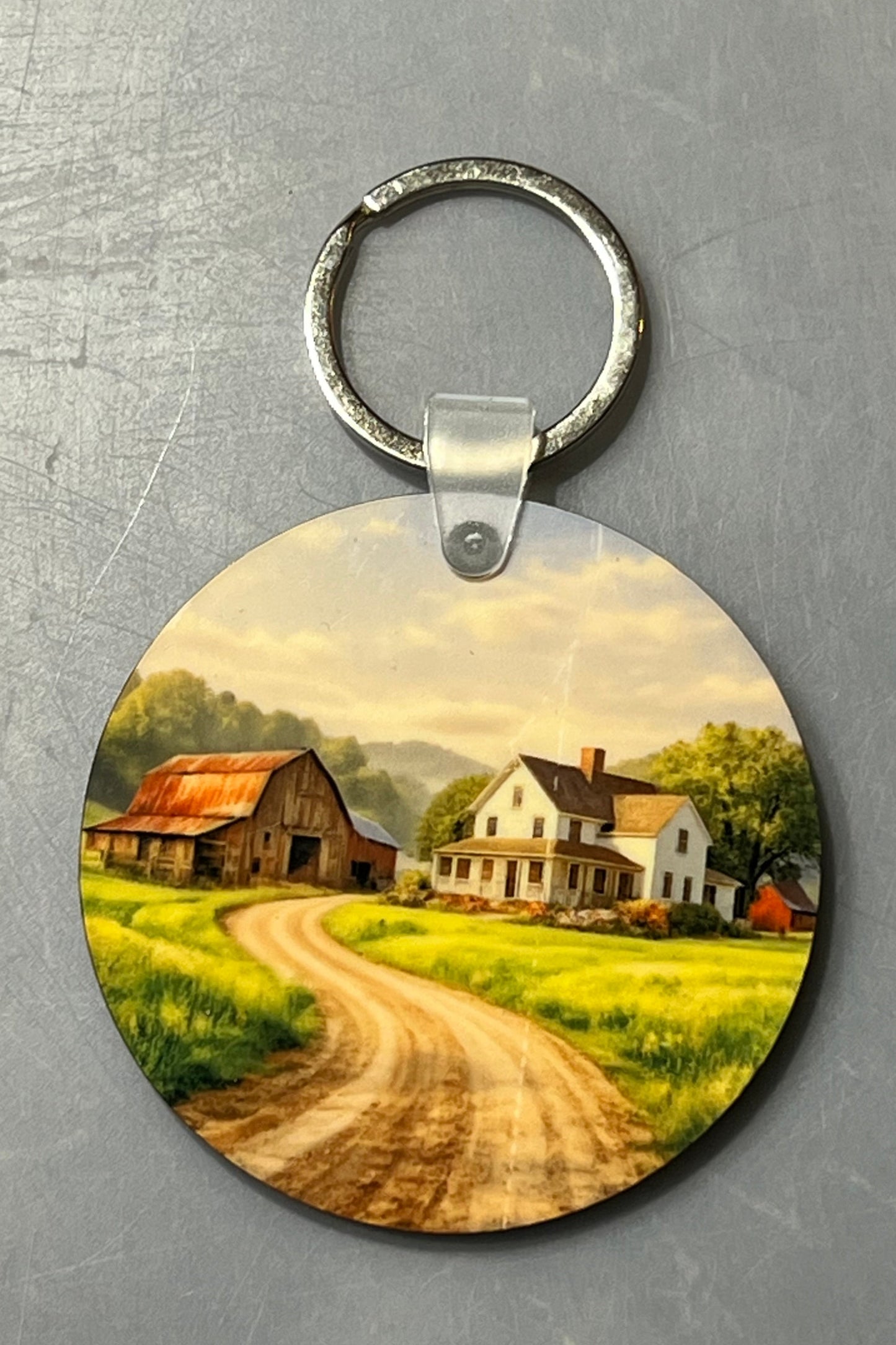 A round wood keychain featuring a printed scenic landscape of a rural farm with a dirt road, barns, and houses under a soft sky, shown on a textured gray background.