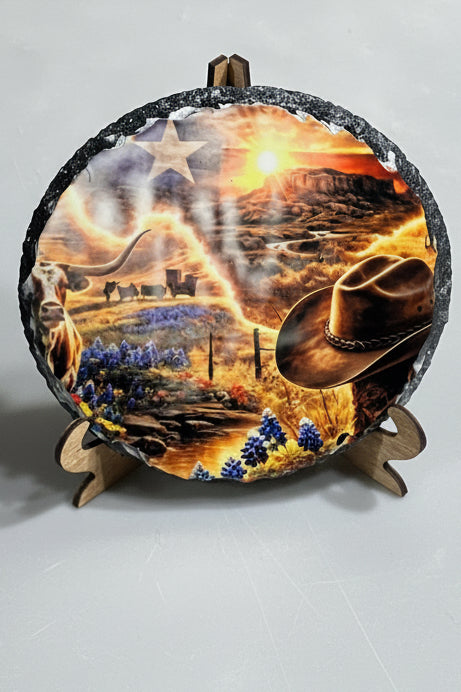 A round decorative slate plaque with a rustic black textured rim, displayed on a small wooden stand. The plaque features a detailed western landscape illustration with a cowboy hat, longhorn cattle, bluebonnets, a sunset, and a star.