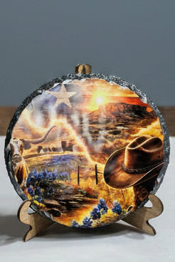A circular decorative plaque featuring a colorful western landscape illustration with a cowboy hat, longhorn cow, blue flowers, and a sunset, displayed on a small wooden stand.