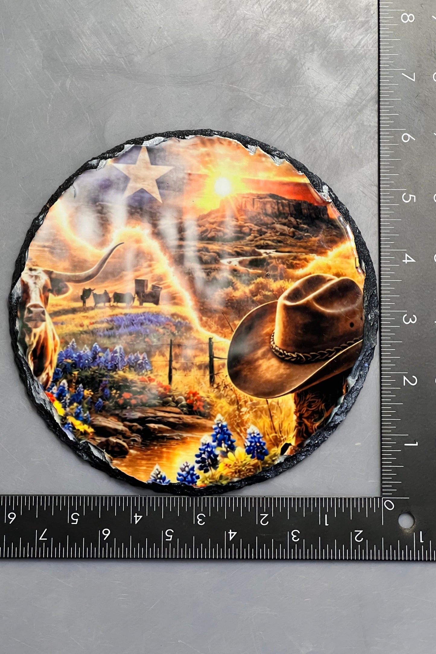 A round slate plaque with a western-themed design, shown next to black metal rulers to indicate its size. The plaque features a cowboy hat, a cowboy boot, a star, and a desert canyon scene.