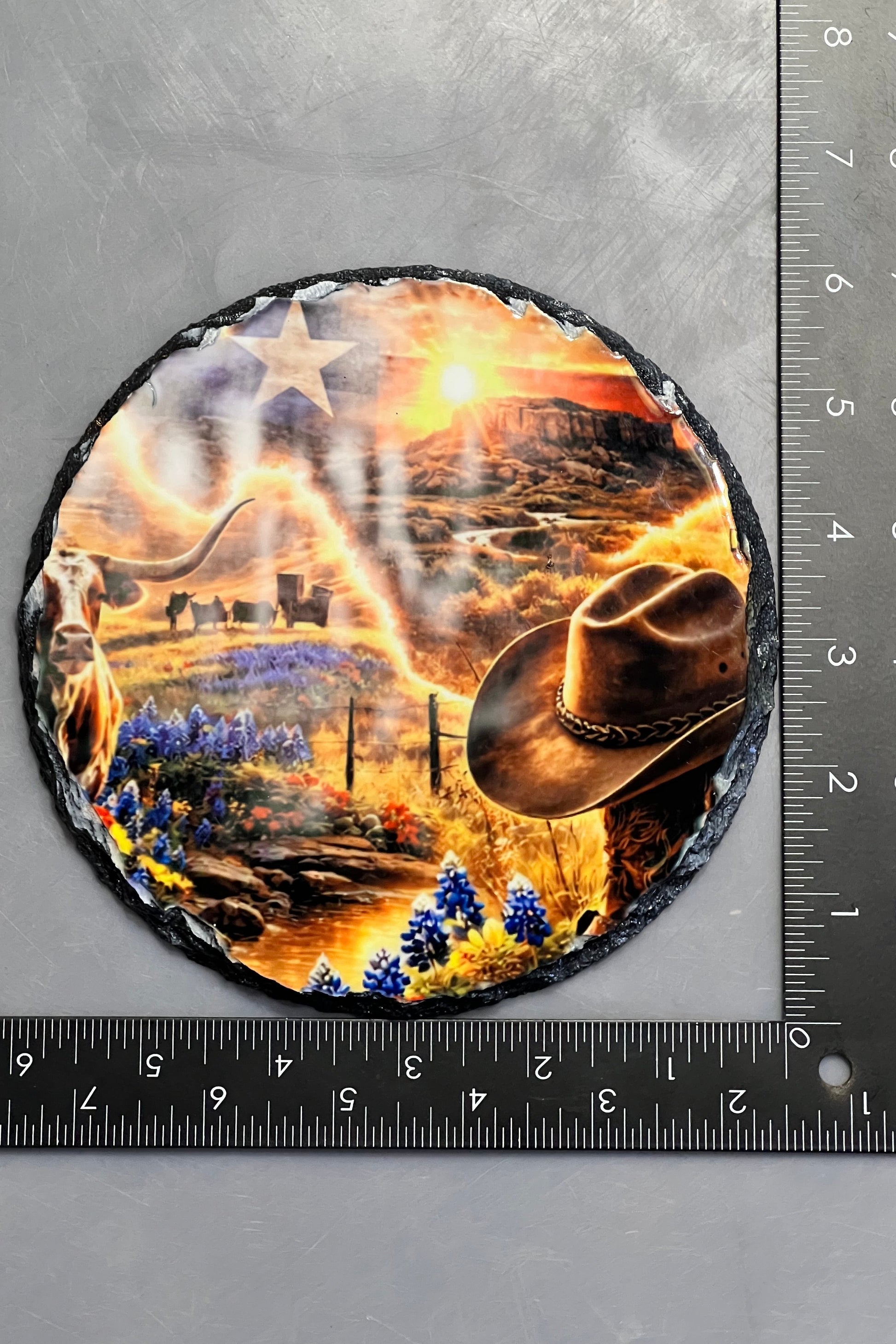 A round slate plaque with a western-themed design, shown next to black metal rulers to indicate its size. The plaque features a cowboy hat, a cowboy boot, a star, and a desert canyon scene.