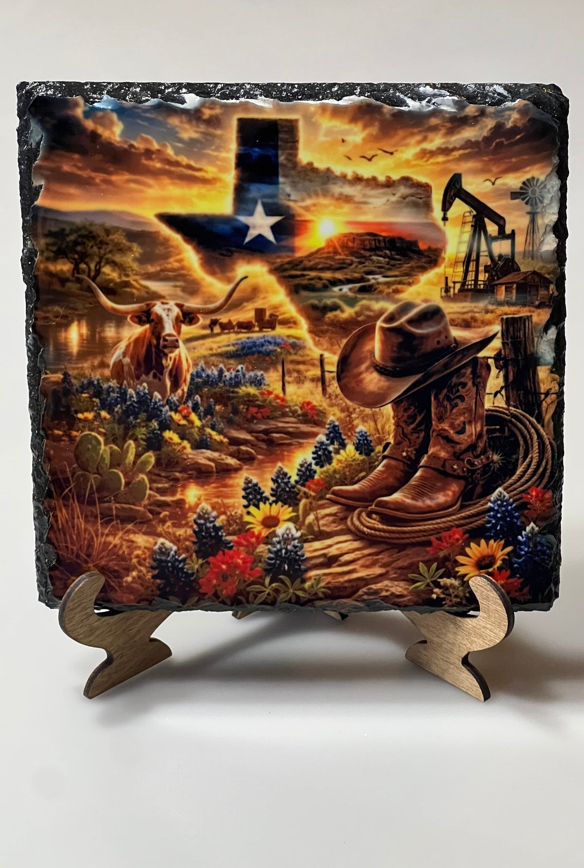 A square decorative slate plaque featuring a detailed Texas-themed cowboy landscape scene with a longhorn, boots, hat, pumpjack, flowers, and a Texas-shaped state outline at sunset, displayed on a small wooden stand against a plain light background.