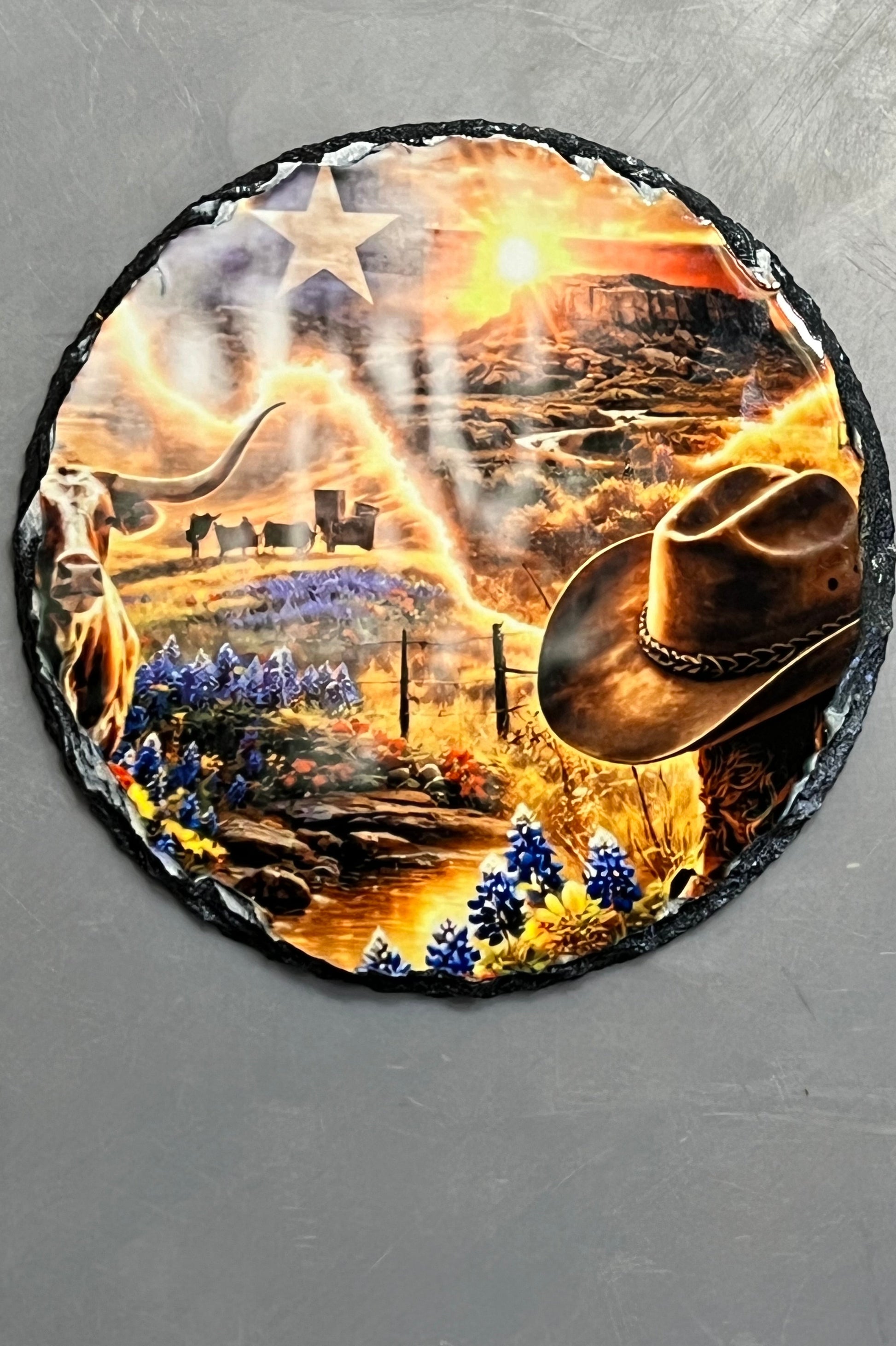 A circular piece of slate featuring a western-themed design with a cowboy hat, stars, mountains, and a bright sunburst, photographed on a textured gray background.
