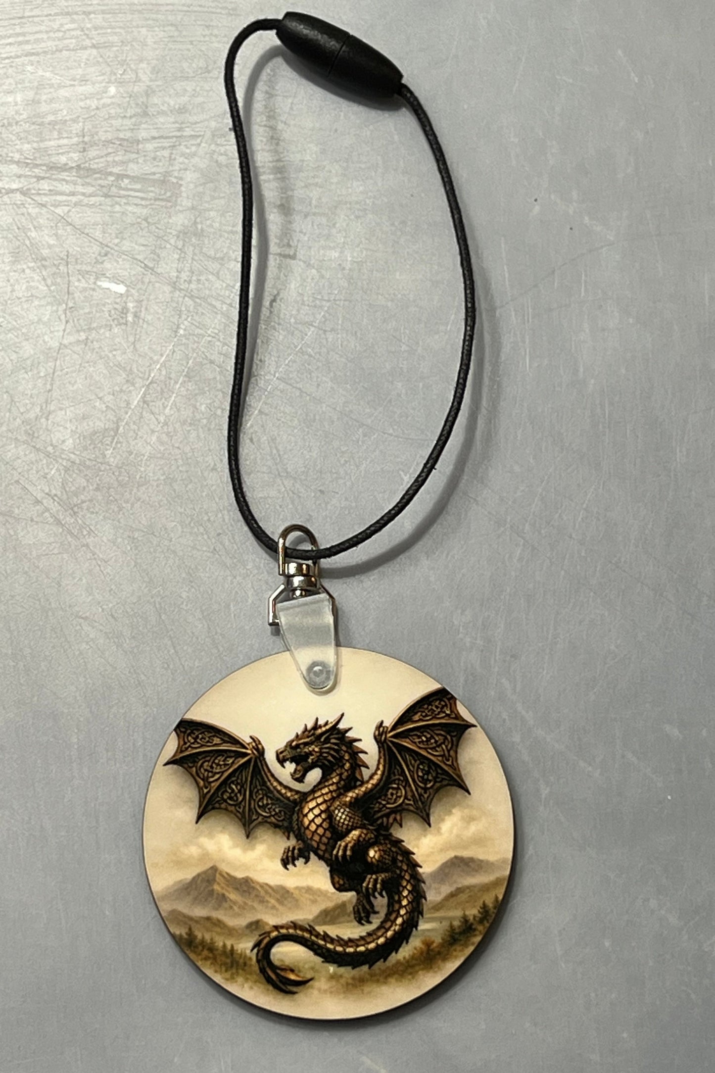 A circular hanging decor featuring a detailed illustration of a dark brown dragon over a beige and gray mountain landscape, suspended from a black cord on a textured gray background.