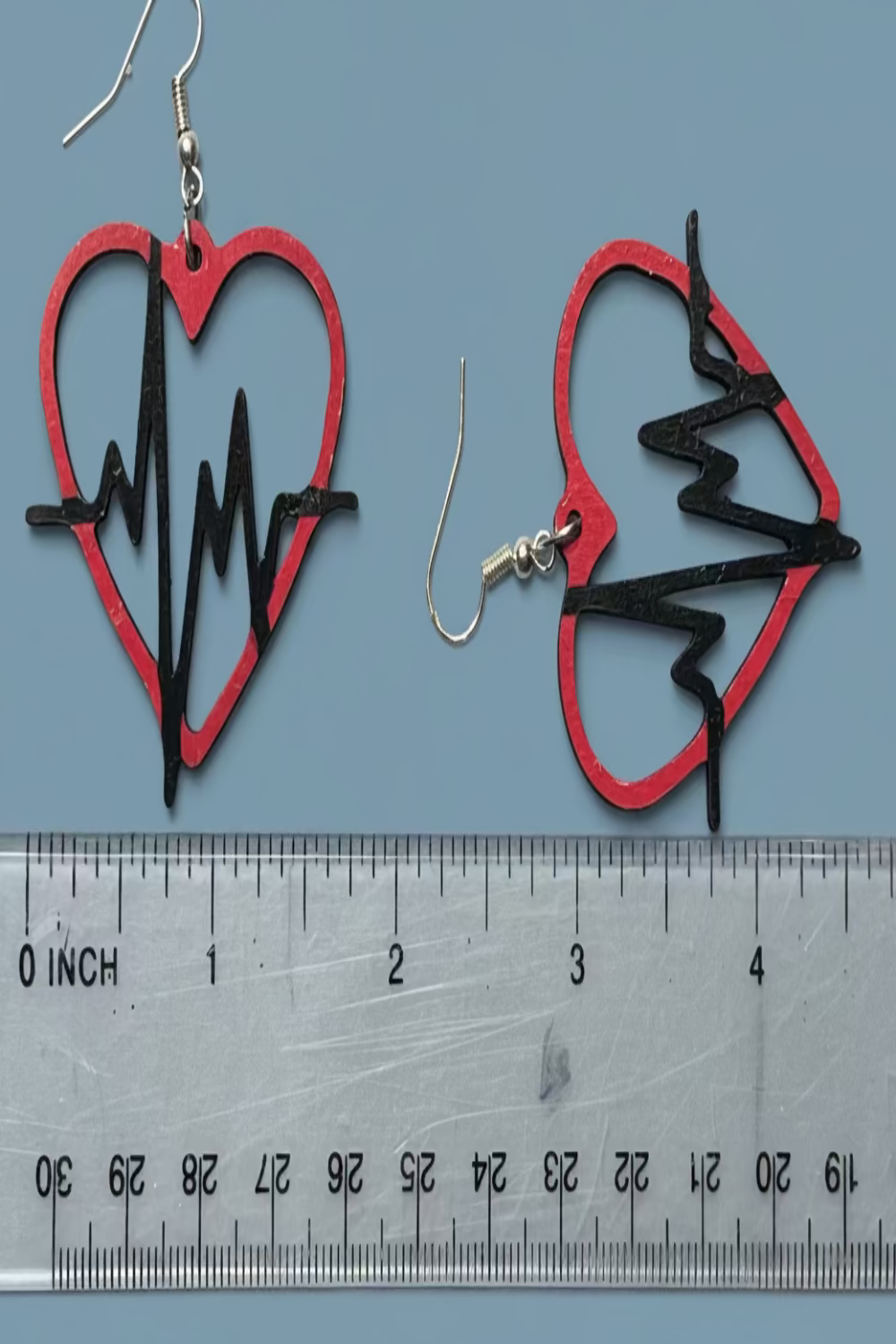 Heart-shaped earrings with a black heartbeat line design next to a ruler on a blue background