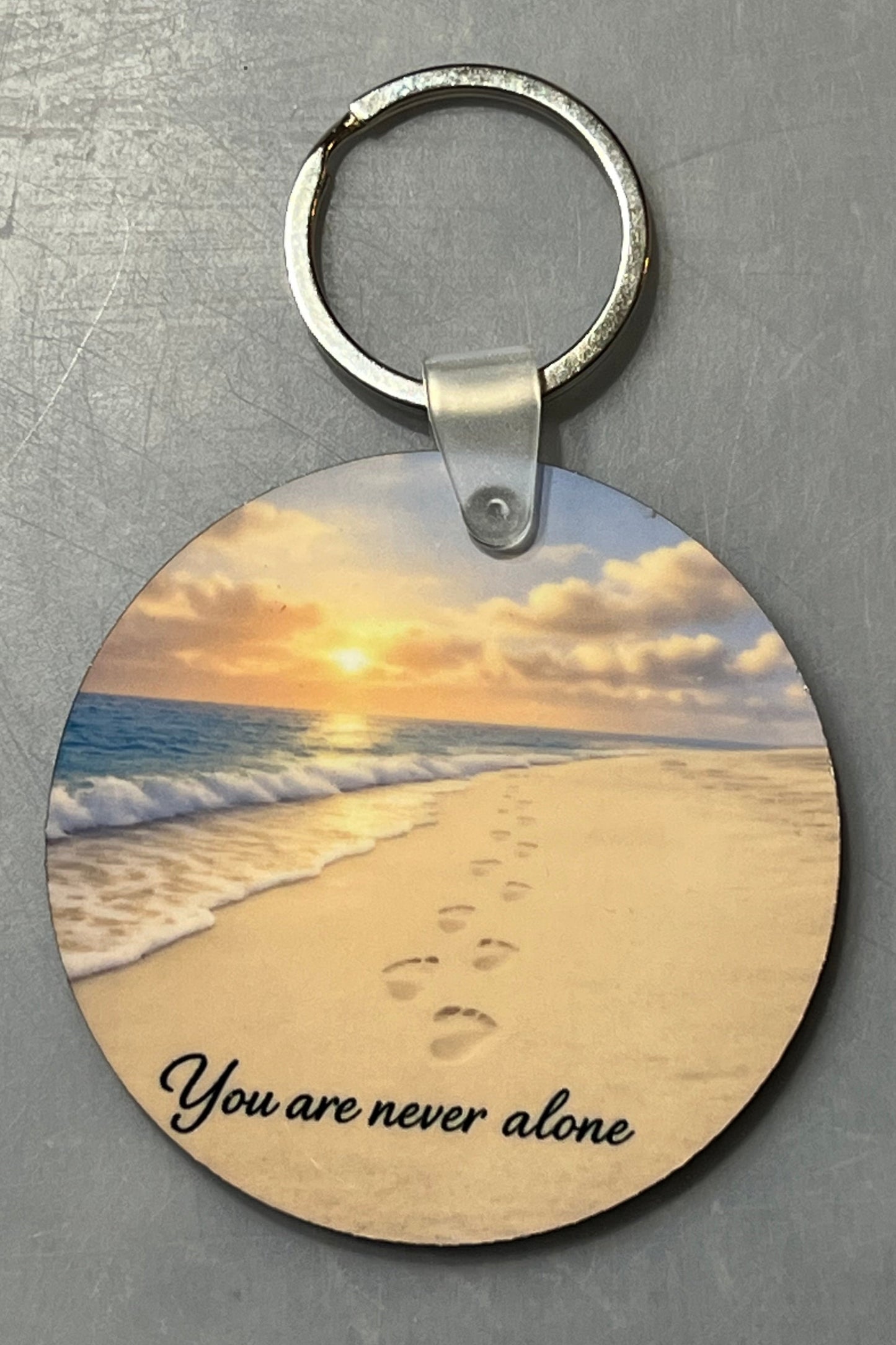A round wood keychain featuring a printed beach sunset scene with footprints in the sand and the text 'You are never alone' at the bottom, attached to a silver split ring and photographed on a gray textured background.